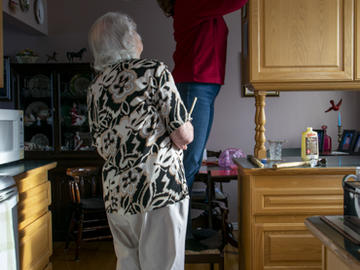 A middle-aged woman stands on a chair to fix or adjust something at the top of a kitchen cupboard, while an elderly woman looks on