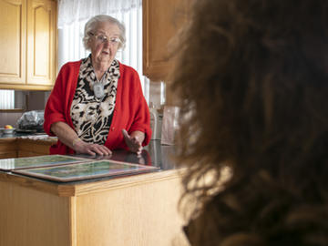An elderly woman stands in a kitchen and talks to a middle-aged woman, whose back is to the camera