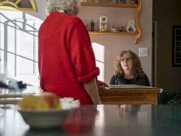 A middle-aged woman sits at a table talking to an elderly woman, who is standing up,  back to the camera