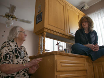 A middle-aged woman sits cross-legged on a kitchen counter, talking to an elderly woman, who is seated on a chair next to the counter.