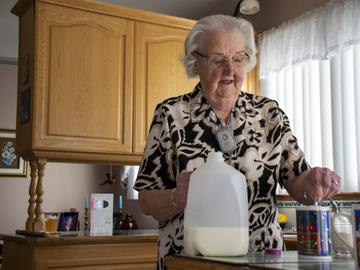 An elderly woman stirs milk into a Christmas mug. A medic alert button is visible on a cord around her neck.