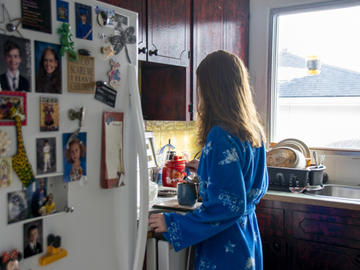 A young woman wearing a robe and holding a mug stands facing the fridge and stove, her back to the camera
