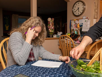 A woman reads a printed paper at a kitchen table, pen in hand, while in the foreground, someone serves themselves salad