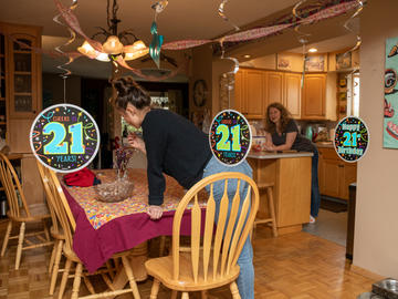 Two people decorate a kitchen for a party. There are streamers and banners reading "Cheers 21 years!"
