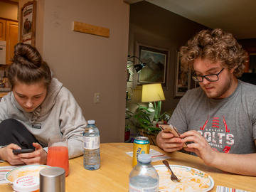Two young adults sit at a table together after a meal. They are both looking at their phones.