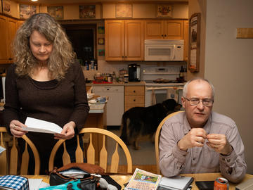 A middle aged couple at their table. The man looks pensive and stares into the distance. The woman stands beside him reading a piece of paper. Their dog is visible in the kitchen behind them.