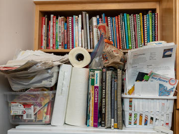 The top of a fridge is cluttered with recipe books, rubbermaid bins and buckets, papers and paper towel