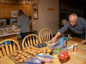 Two people are doing food prep in their kitchen. Two dozen muffins are cooling on a rack on a table, which also holds multiple ziploc bags of prepped food.