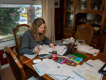 A woman sits at a dining room table cluttered with financial statements and highlighter pens. There is an adding machine beside her.