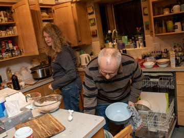 Two people work in a kitchen together. The cupboard doors are open.