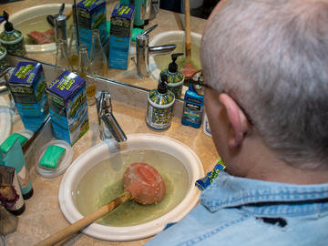 A man works on a plugged sink. He has a plunger and a box of liquid plumbing solution.