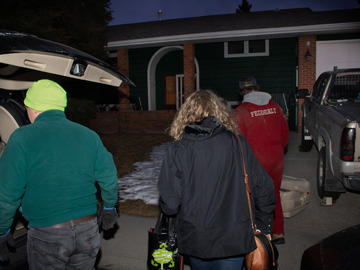 Three people, dressed for evening winter weather, walk towards their house. The back of their van is open.