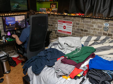 A young man plays video games in his room. Folded laundry on his bed waits to be put away.