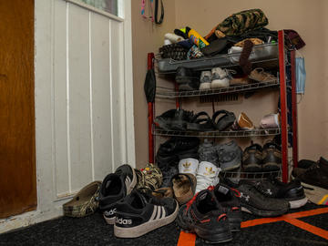 A four shelf shoe rack groans under the weight of many pairs of shoes. Ten or so more pairs are strewn on the floor in front of the shelf.