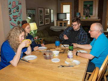 A family of four adults sits at a table with pancakes and syrup. The elder male is speaking and the younger male looks at his phone