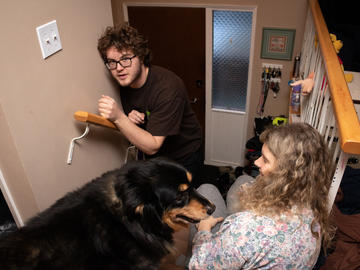 A woman and dog sit on the front stairs of a house while a younger man tries to squeeze up the stairs past them