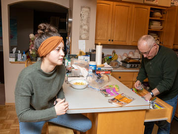 A young woman sits at a kitchen island while a middle-aged man rummages in its drawers