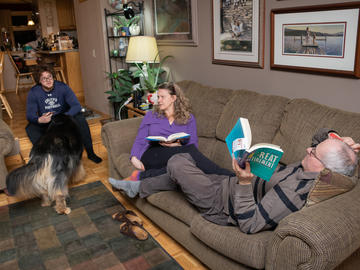 Three people and a dog chat in a living room. The older adults sit together on a couch with open books in their hands