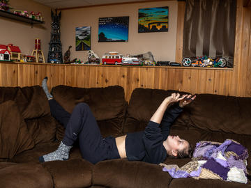 A young woman stretches up her arms while laying on a big sectional couch in a rec room. Her phone is in her hands and she is gesturing. There is a train set and cars on the wall behind her.