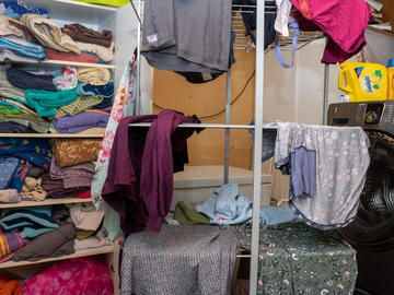 A family's cluttered laundry room with clothes hanging on a drying rack and a full shelf of linens