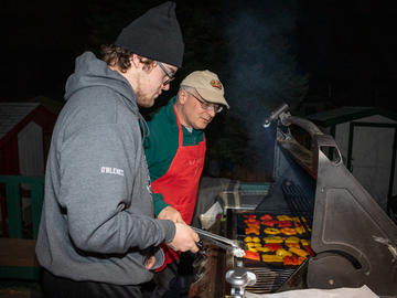 A younger and older male grill peppers on the barbecue. It's night time, and the younger man wears a hoodie and toque, while the older man wears an apron over a hoodie and a ball cap.
