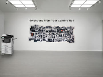 On a gallery wall, the text "selections from your camera roll." A wide selection of images is arranged below the text. There is a photocopier/printer in the room. 