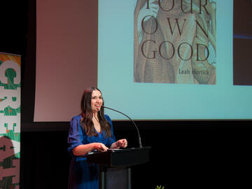 Leah Horlick speaks at a podium during Hello/Goodbye 2022. She wears her dark hair long and a blue dress.