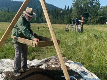 A researcher sifts through dirt using a sifting tool that hangs from a wooden tripod structure.