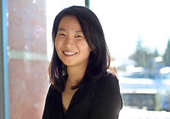 Teresa Wong smiles and looks at the camera. She wears a black shirt, her hair loose, and stands in front of a sun-filled window.
