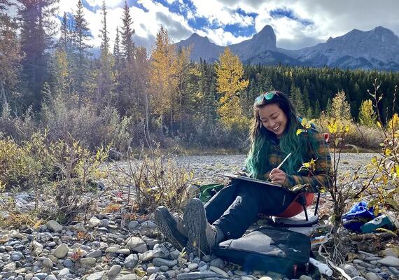 A female student with long turquoise hair sits in front of a natural wetland, writing in her notebook. She is smiling widely at her work.