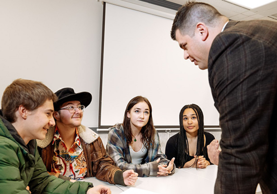 A professor leans over a table to chat with students. They all look interested and engaged.