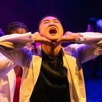 A student in a dance piece tilts back her head in a yell or scream. Her hands cup her chin.