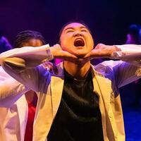 A student in a dance piece tilts back her head in a yell or scream. Her hands cup her chin.