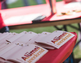A stack of red and gold Alumni Weekend programs sits on a table
