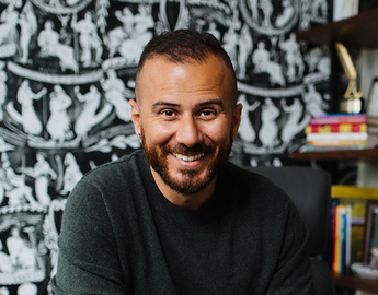 Danny Ramadan smiles in front of a bookcase and graphical wallpaper.